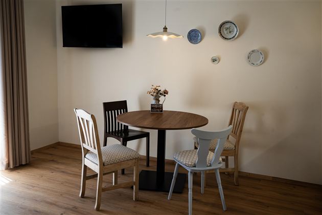Een gezellige eettafel van hout met drie stoelen in een lichte kamer. Aan de muur hangen enkele borden en een televisie is zichtbaar.