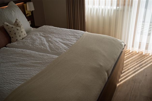 Een gezellige slaapkamer met een groot bed en zachte dekens. Daglicht stroomt door de gordijnen en werpt interessante schaduwen op de vloer.
