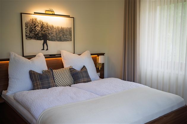 Een gezellige slaapkamer met een groot bed en verschillende kussens. Op de achtergrond hangt een afbeelding van een sneeuwbos.