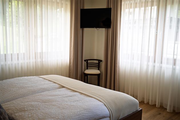 Een lichte slaapkamer met een groot bed en een televisie aan de muur. Naast het bed staat een enkele stoel en grote, lichte gordijnen laten het licht binnen.