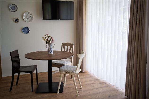 Een gezellige eetruimte met een ronde tafel en drie stoelen. Aan de muur hangen decoratieve borden, en grote ramen laten veel licht binnen.