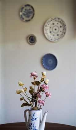Een elegante kan met kleurrijke bloemen staat op een tafel. Op de achtergrond hangen decoratieve borden aan de muur.