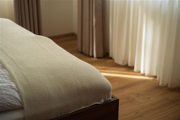 Een gezellige slaapkamer met een bed en een lichte, neutrale deken. Zachte gordijnen laten warm licht binnen.