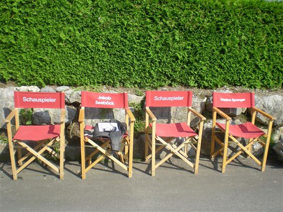 Vier Regiestühle mit roten Sitzflächen stehen auf dem Boden. Neben jedem Stuhl steht der Schriftzug "Schauspieler".
