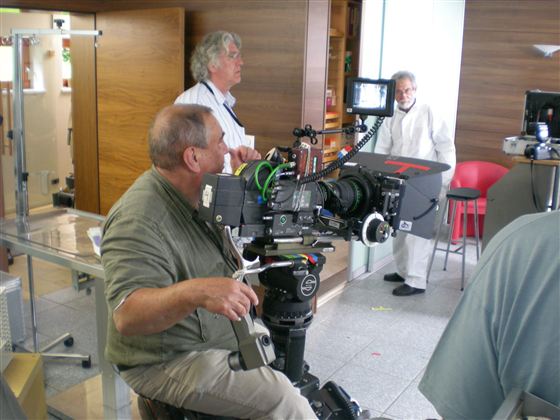 Ein Kameramann sitzt vor einer großen Kamera. Im Hintergrund sind weitere Personen und ein modernes Raumambiente zu sehen.