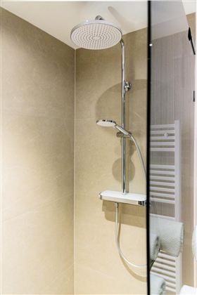 A modern shower with a large rain shower head and a clear, elegant design. In the background, bright tiles and a towel holder are visible.