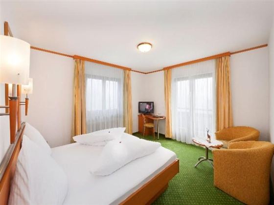 Ein gemütliches Hotelzimmer mit einem Doppelbett und einem kleinen Tisch. Große Fenster lassen viel Licht hinein und die grüne Teppichfläche sorgt für eine angenehme Atmosphäre.