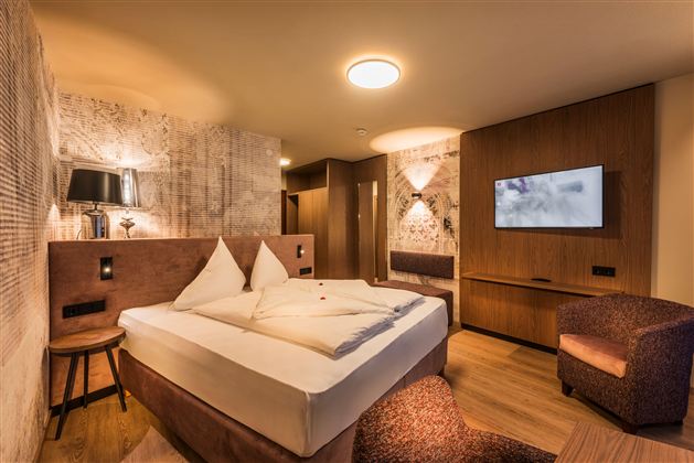 Ein modernes Hotelzimmer mit einem großen Bett, eleganter Holzverkleidung und einem Fernseher an der Wand. Gemütliche Möbel und warmes Licht sorgen für eine einladende Atmosphäre.