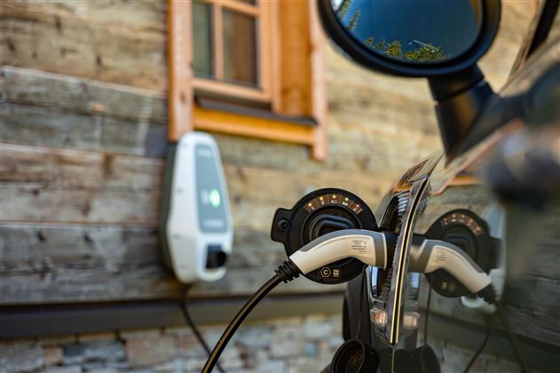 Ein Elektroauto wird an einer Ladestation aufgeladen. Im Hintergrund ist ein Holzhaus mit einem Fenster zu sehen. 
Chalet Heualm Kelchsau Kitzbüheler Alpen