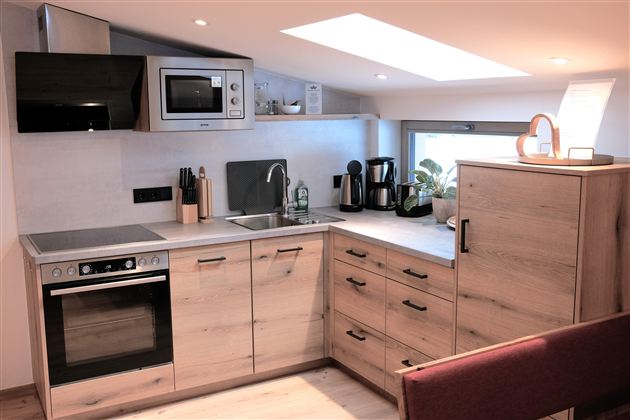 A modern kitchen with wooden surfaces and new appliances. It features a stove, a sink, and a microwave, as well as a view to the outside.