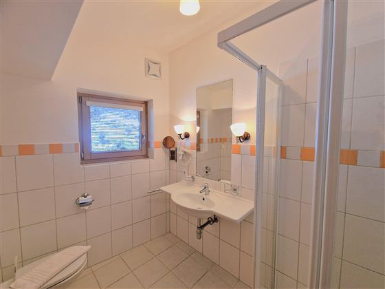 A bright bathroom with a shower, sink, and mirror. It has white tiles and a window that lets in light.