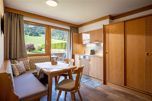 Ein gemütliches Wohnzimmer mit Holzmöbeln und einem Tisch. Durch das Fenster sieht man eine grüne Landschaft mit Bergen.