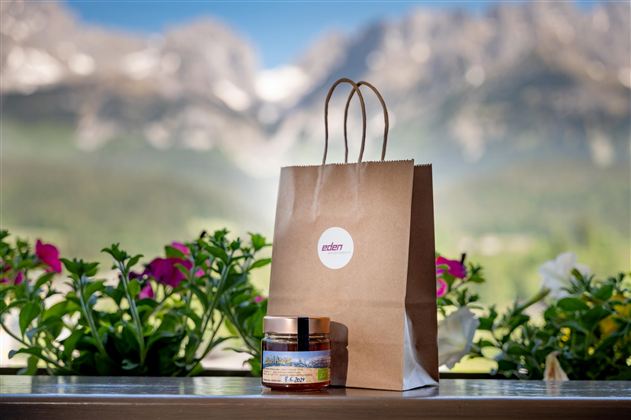 Eine braune Papiertüte steht neben einem Glas mit einer Creme. Im Hintergrund sind blühende Pflanzen und Berge zu sehen.