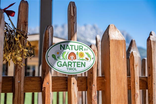 Ein Schild an einem Holzzaun mit der Aufschrift "NATUR GARTEN TIROL". Im Hintergrund sind Berge und ein klarer blauer Himmel zu sehen.