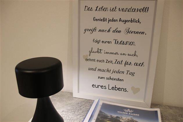 Ein stilvolles Schwarz mit einer runden Lampe und einem gerahmten Zitat über das Leben. Darunter liegt ein Prospekt mit einer schönen Landschaft.