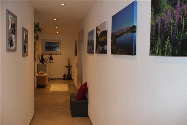 Ein schmaler Flur mit weißen Wänden und mehreren Bildern an der Wand. Am Ende des Flurs gibt es einen kleinen Tisch und eine Pflanze.