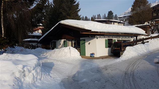 Een gezellig chalet, omringd door veel sneeuw en bomen. De zon schijnt op het dak, dat bedekt is met een laag sneeuw.