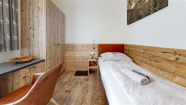 A simple room with wooden paneling and a single bed. There is a desk, a chair, and a window with curtains.