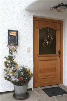 Een versierde houten voordeur met een krans. Naast de deur staat een kleine kerstboom in een pot.