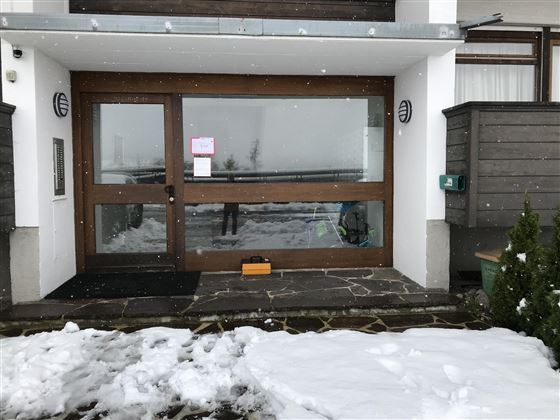 An entrance with large windows and a wooden porch. Outside, there is snow and it is lightly snowing.