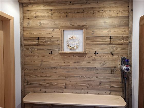 Eine helle Holzwand mit einer zentralen Wanddekoration und mehreren Haken. Darunter befindet sich eine einfache Holzbank.
Akkustaubsauger vorhanden