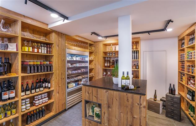 Een uitnodigende verkoopruimte met houten planken vol flessen en delicatessen. In het midden staat een verkoopbalie met enkele wijnflessen.