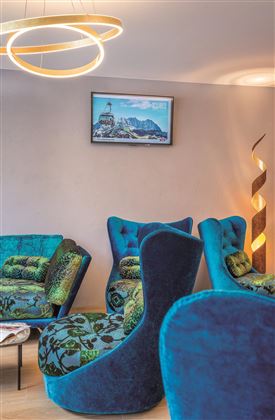 Een moderne woonkamer met meerdere blauwe fauteuils en een unieke kroonluchter. Aan de muur hangt een schilderij van bergen en een stad.