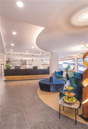 Een moderne ontvangsthal met elegante meubels en lichte muren. Op de voorgrond staat een ronde tafel met fruit en comfortabele stoelen.