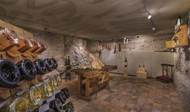 Een gezellige wijnkelder met natuurstenen muren en warm licht. Op de planken zijn flessen en kaas te zien, en in het midden staat een grote houten tafel.