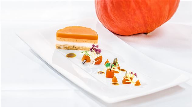 Een kunstzinnig opgemaakt dessert met lagen van pompoen en een decoratie van kleurrijke groenten. Op de achtergrond ligt een grote pompoen.