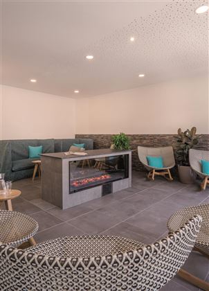 Een moderne ontspanningsruimte met comfortabele zitplaatsen en een centrale open haard. De muren zijn in neutrale kleuren gehouden, aangevuld met groene planten.