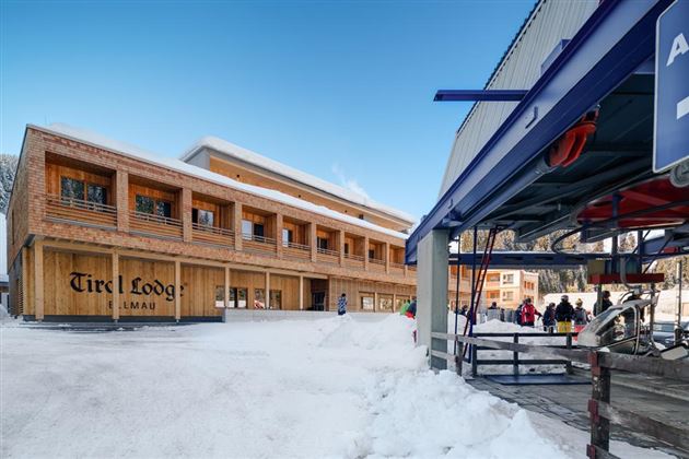 A modern wooden building in the snow with the inscription "Tire Lodge". In the foreground, a ski lift and some visitors can be seen.