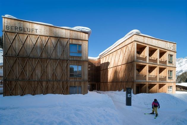 A modern wooden building in a snowy landscape. In the foreground, a skier can be seen heading towards the accommodation.