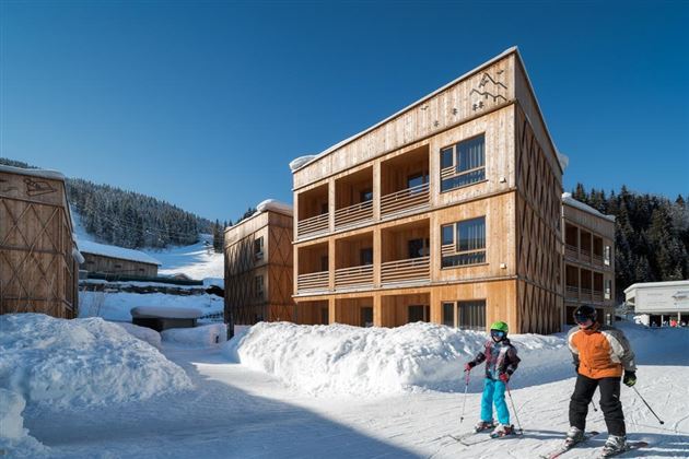 A wooden facade of the building in the snow, surrounded by snow-covered mountains. Two skiers are standing on the path and enjoying the surroundings.