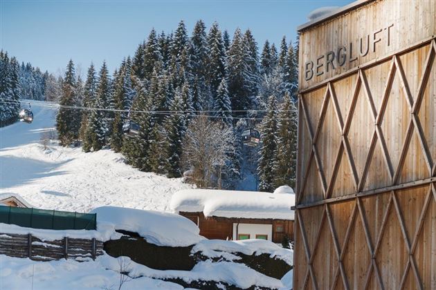 A snowy mountain landscape with dense coniferous trees and a ski lift in the background. In the foreground, there is a wooden building with the inscription "BERGLUFT".