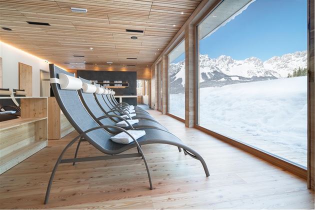A modern relaxation room with loungers and large windows that offer a stunning view of snow-covered mountains. The bright wood and cozy atmosphere invite relaxation.