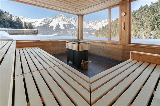 A cozy sauna area with wooden benches and a fireplace. In the background, snow-covered mountains stretch under a blue sky.