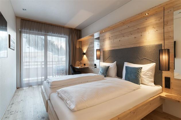 A modern bedroom with two beds and stylish wooden decor. Large windows provide plenty of daylight and a view outside.