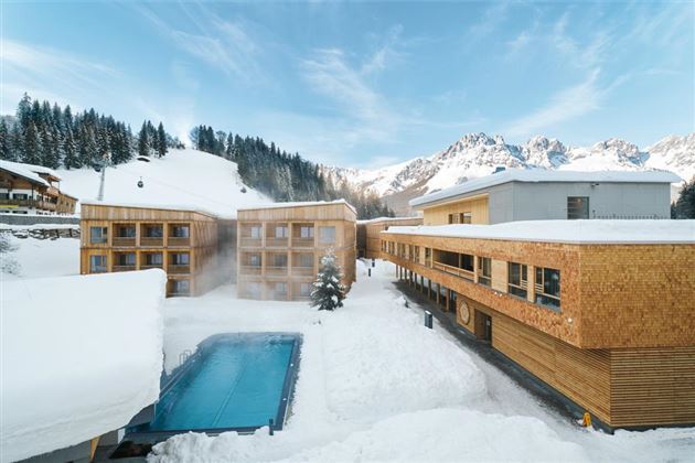 A modern hotel building in a snow-covered landscape. In the foreground, there is a heated pool, and in the background, snow-capped mountains can be seen.