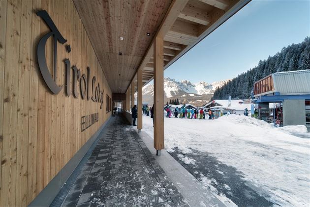 A modern entrance area with wood paneling in a snow-covered environment. In the background, people can be seen preparing for a ski trip.