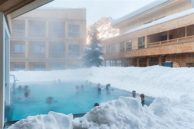 A pool in the middle of snow with several people relaxing in the water. In the background, the wooden buildings of an accommodation can be seen.