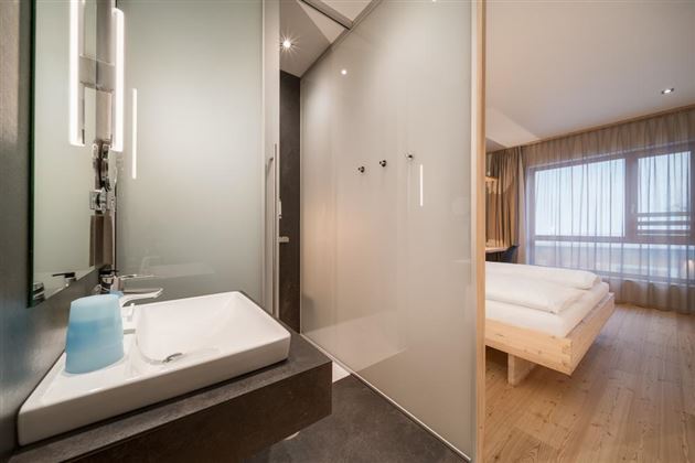 A modern bathroom with a sink and a glass door shower cabin. In the background, there is a bright bedroom with a large window and a wooden bed.