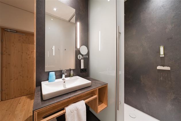 A modern bathroom with an elegant sink unit and a large mirror. The walls are dark and the shower is enclosed with glass.