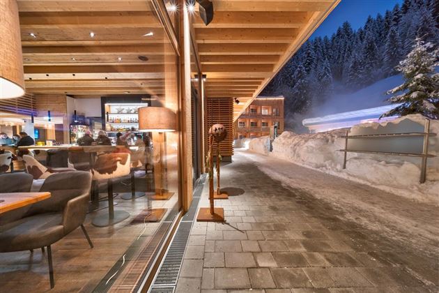 A modern restaurant with wooden decor and a cozy atmosphere. Outside, snow lies on the ground and the surroundings are surrounded by wooded mountains.