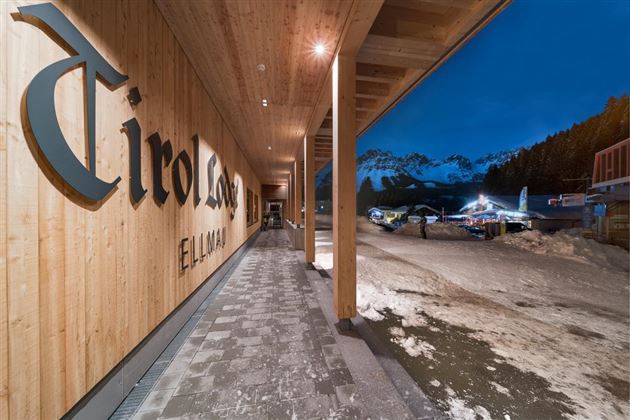 A modern wooden entrance with the inscription "Tirol" and a view of snow-covered mountains. It is dusk, and the surroundings are calm and inviting.