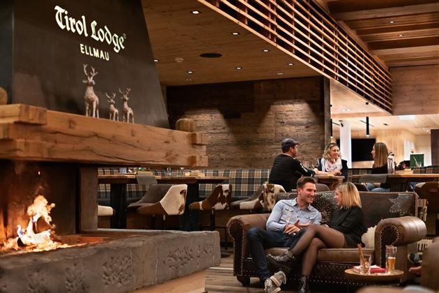 A cozy lounge with an open fireplace and modern wooden furniture. Couples and groups enjoy the relaxed atmosphere at Tirol Lodge Ellmau.