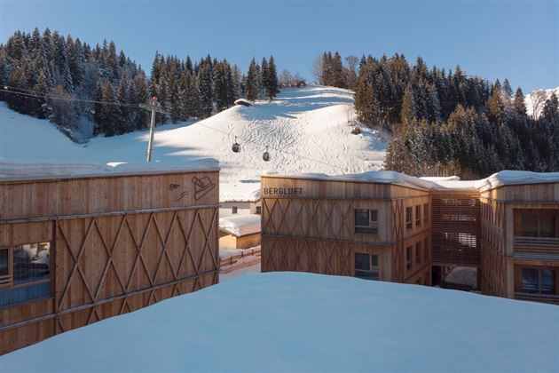 A modern wooden building in the mountains, surrounded by snow-covered hills. In the background, ski lifts and a snowy slope are visible.