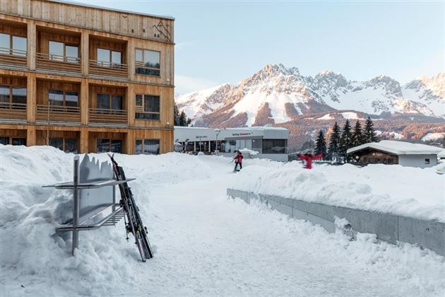 A picturesque winter resort with snow-covered mountains in the background. In the foreground, there is ski equipment and people walking in the snow.