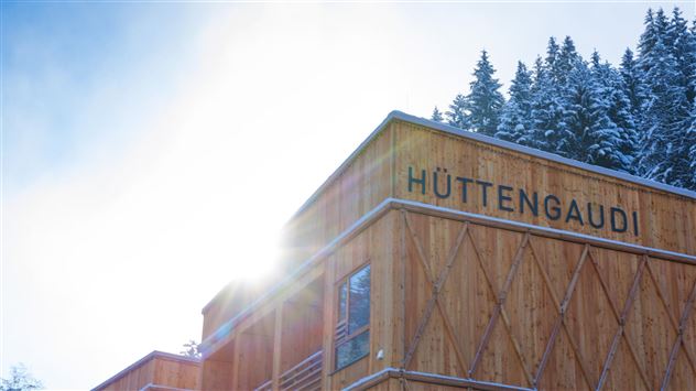 A modern wooden house in the mountains, surrounded by snow-covered fir trees. The sun shines bright and reflects off the building.