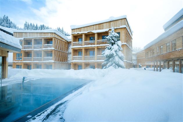 A modern hotel in a snowy landscape. The exterior features a pool, surrounded by snow and wooden architecture.